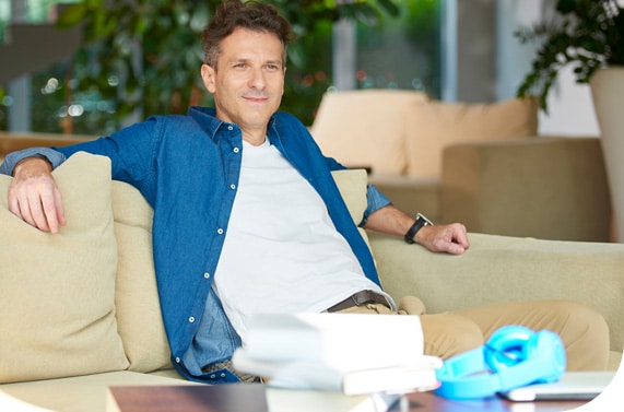 A man wearing a blue shirt over a white t-shirt sits in a casual corporate environment on a beige sofa and smiles