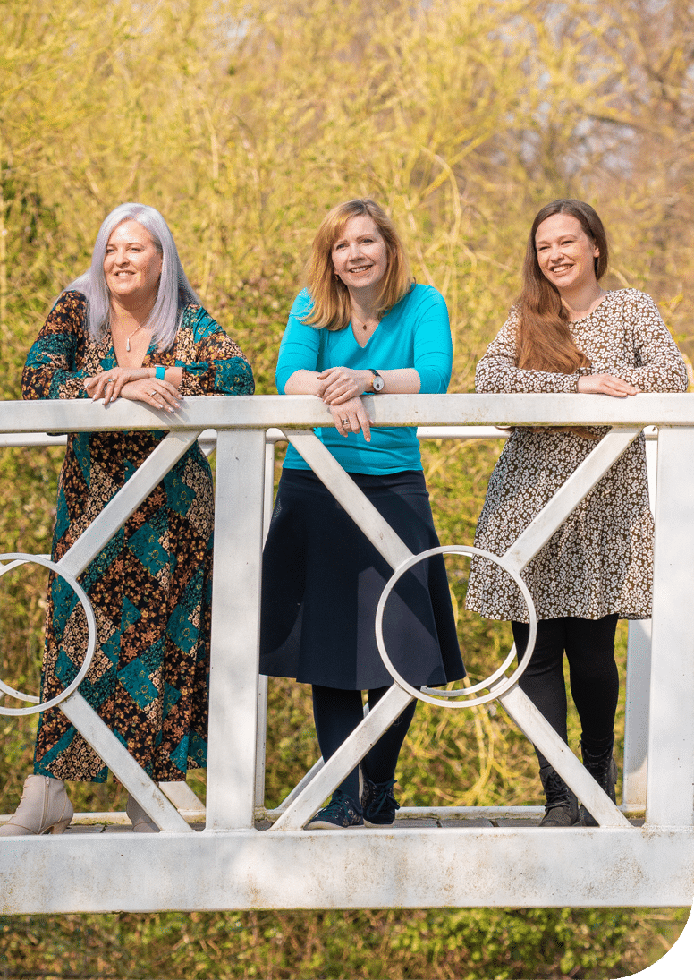 Eleanor, Sarah and Hannah of face2faceHR stand on a bridge in a park on a sunny day smiling