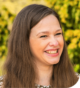 A headshot of Hannah Ambler, grinning with trees in the background