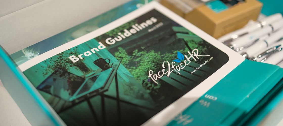 Two women in office wear sit at a desk in conversation. The first holds a sheet of paper above a ringbinder folder.
