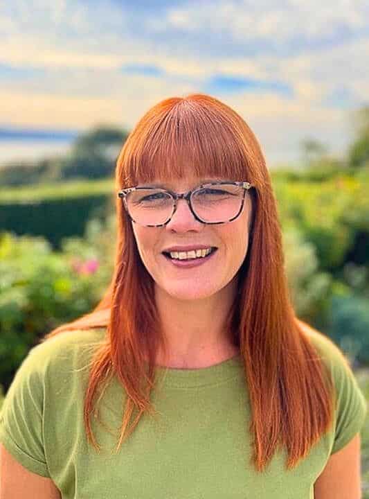 Two women in office wear sit at a desk in conversation. The first holds a sheet of paper above a ringbinder folder.