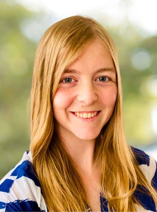 Two women in office wear sit at a desk in conversation. The first holds a sheet of paper above a ringbinder folder.