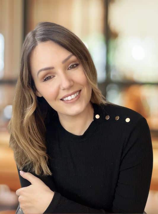 Two women in office wear sit at a desk in conversation. The first holds a sheet of paper above a ringbinder folder.